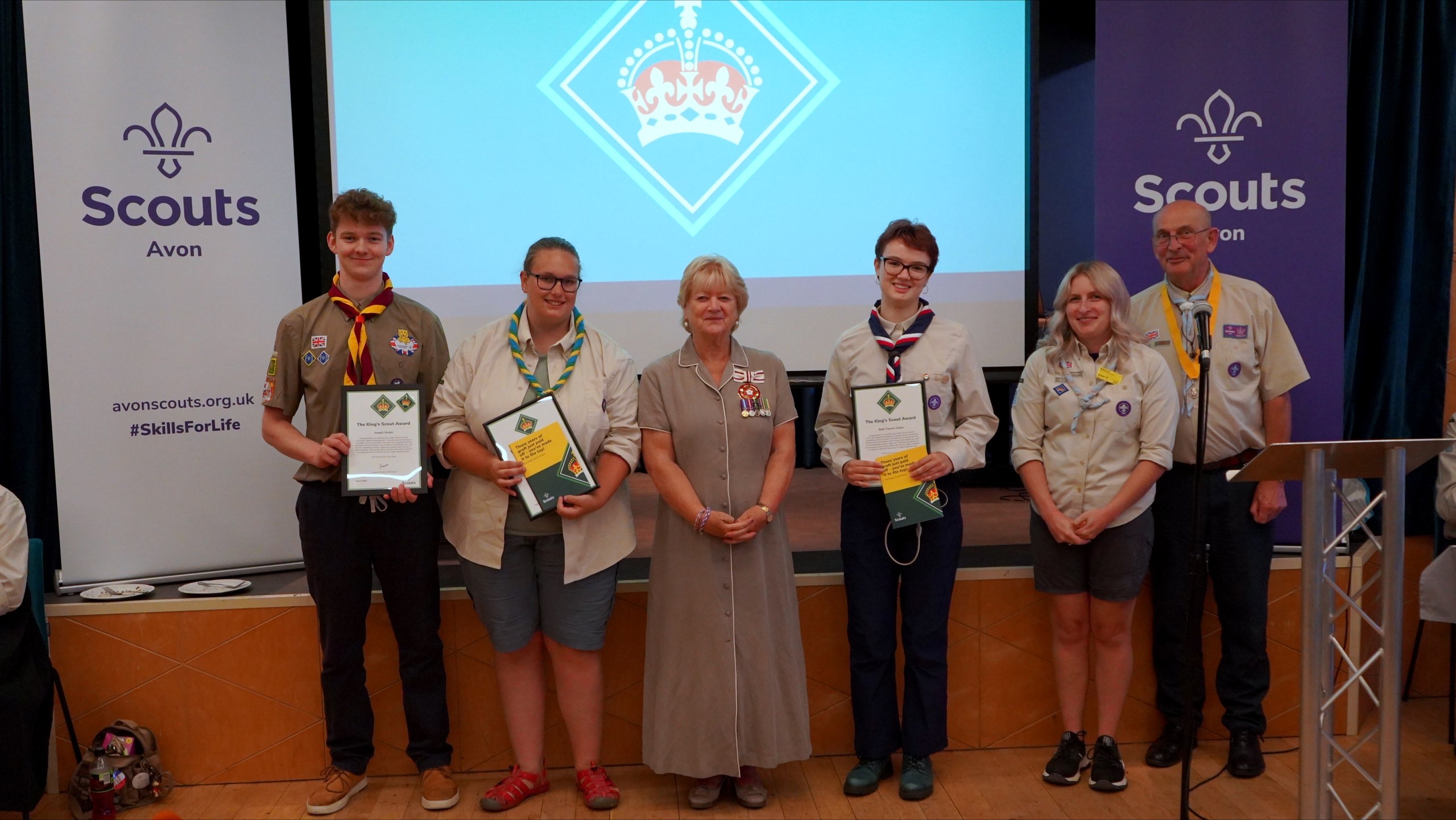 Three King's Scout Award recipients holding their certificates on stage with the guest of honour and two County volunteers, beneath a projected King's Scout Award crown.