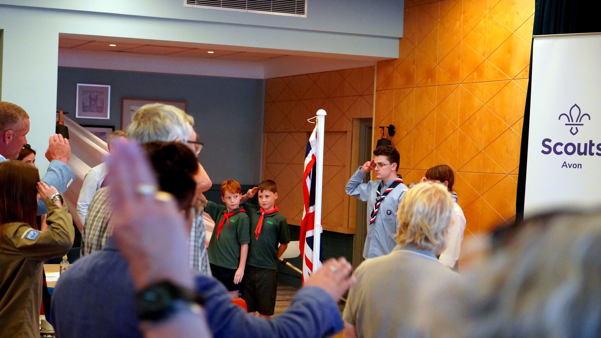 Two Scouts stand either side of the Union flag on its pole while delegates in the hall salute.