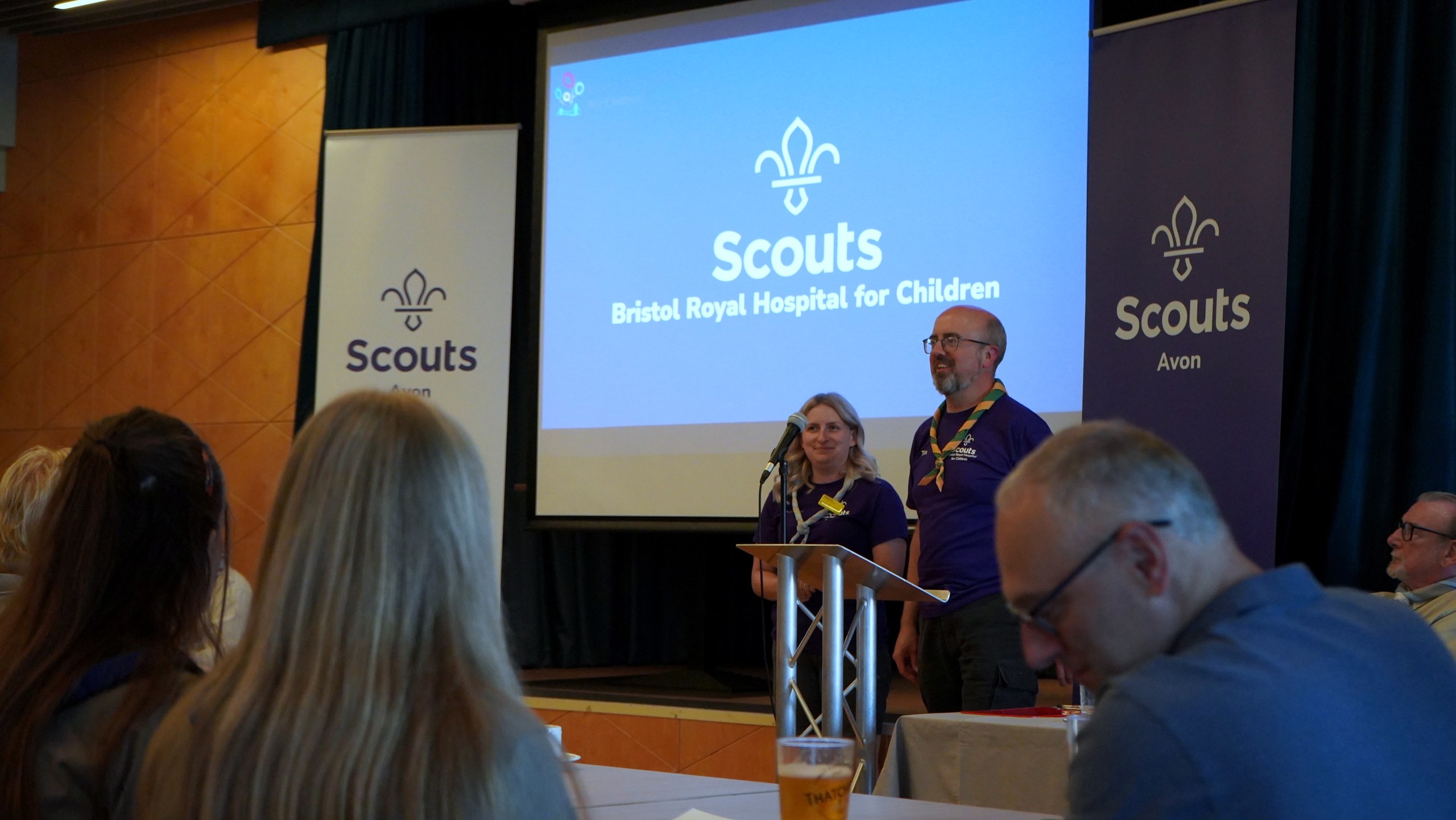 Two volunteers in purple Scouts t-shirts present at a lectern in front of a screen reading 'Scouts — Bristol Royal Hospital for Children'.