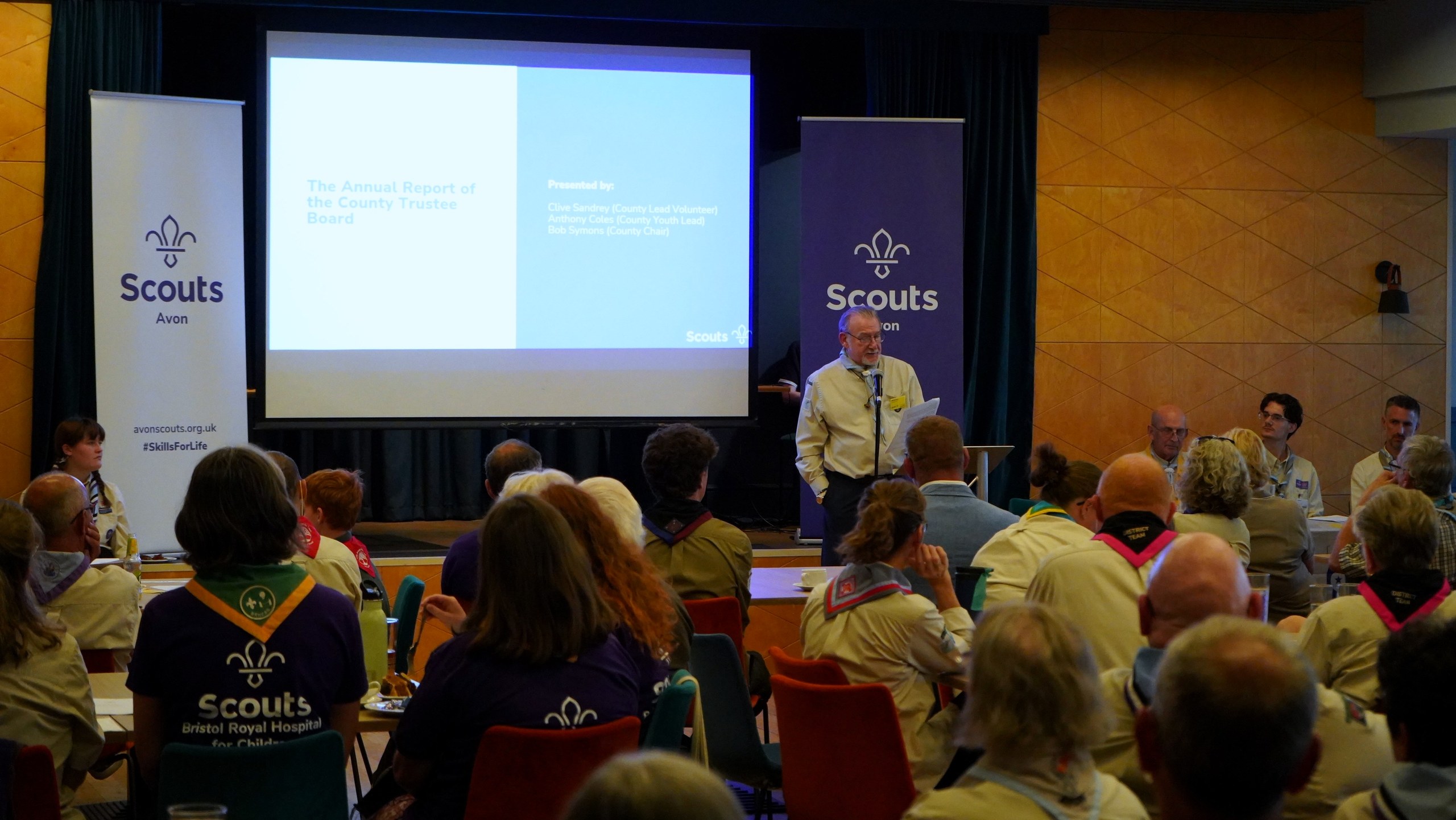 A County volunteer addresses the meeting in front of a screen reading 'The Annual Report of the County Trustee Board'.