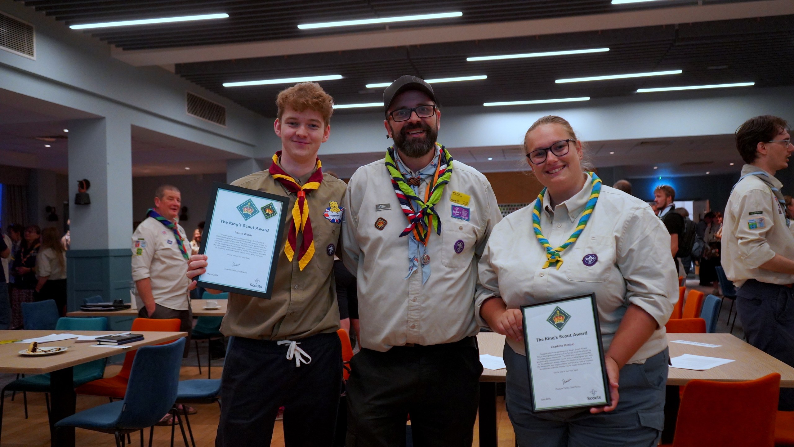 Two King's Scout Award recipients hold their framed certificates either side of a smiling leader in a cap and neckerchief.