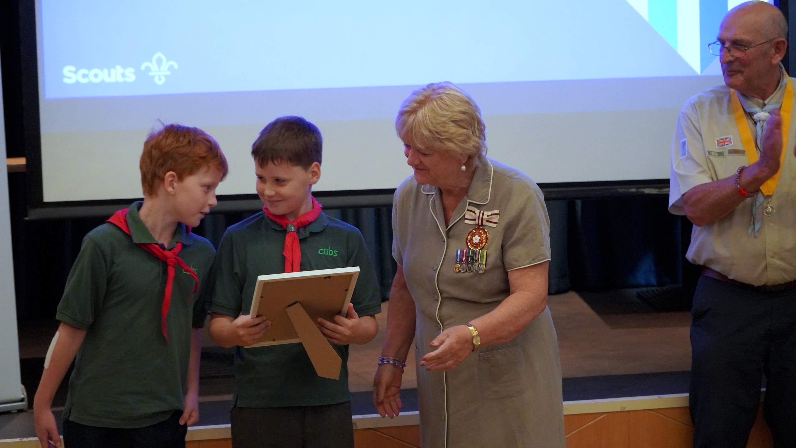 Two Cub Scouts in green tops and red neckerchiefs are presented with a framed certificate by the guest of honour, watched by a County volunteer.