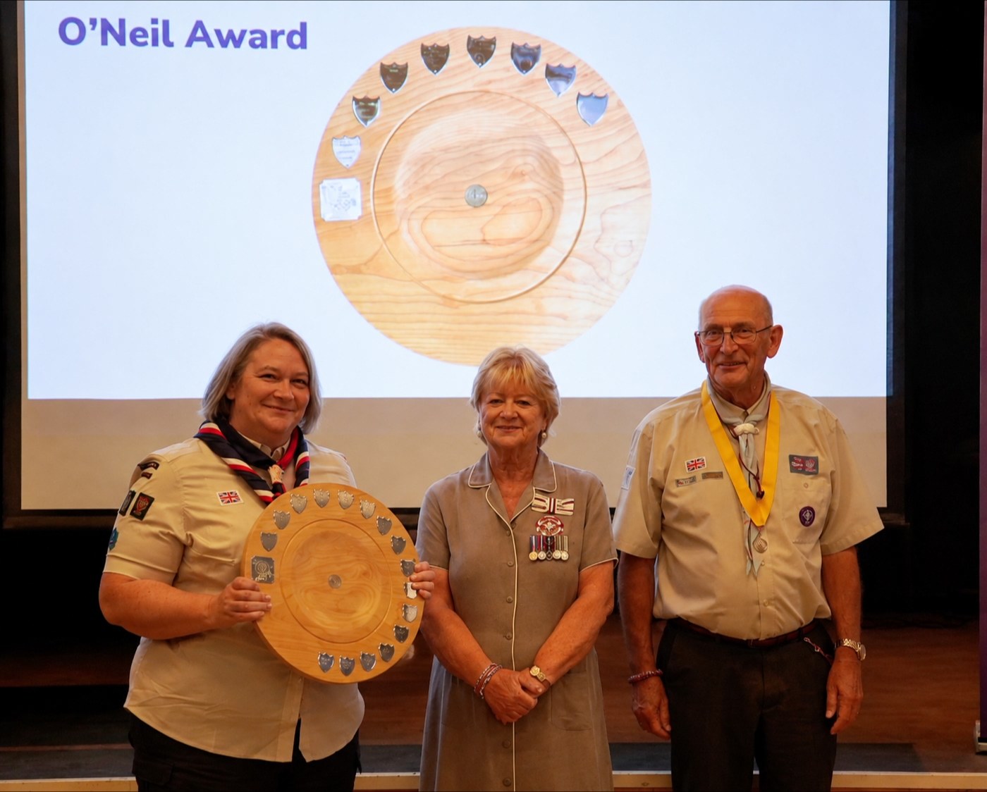A volunteer holds a large wooden shield mounted with engraved shields, standing with the guest of honour and a County volunteer beneath a projected image of the same trophy.