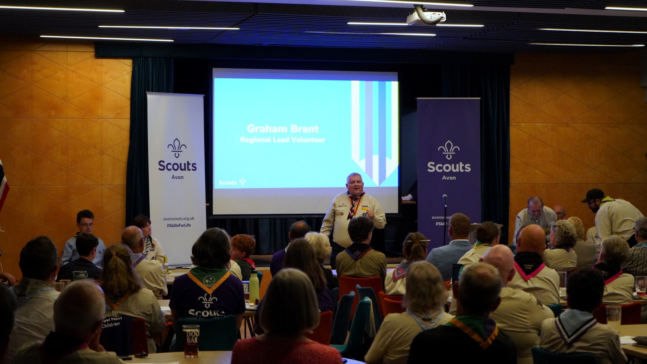 A volunteer addresses the meeting in front of a screen reading 'Graham Brant — Regional Lead Volunteer'.